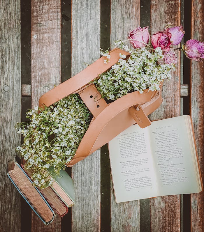 Top view of an open book surrounded by a handbag filled with flowers on a wooden bench.